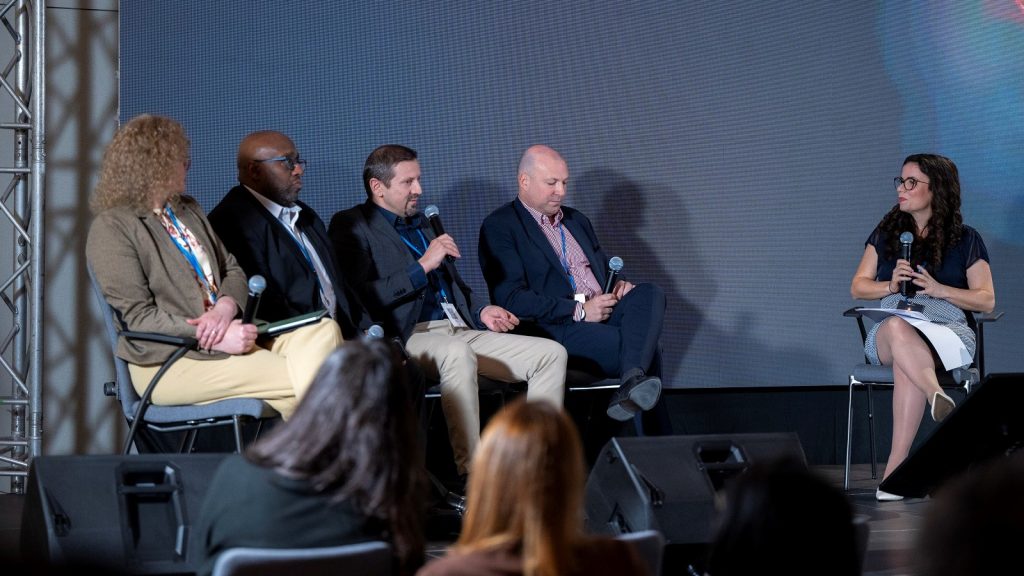 Vielfältiges Panel auf einer Konferenz, mit Diskussionsteilnehmern im Business-Format und einem Moderator, der Fragen stellt, vor einem großen digitalen Bildschirm, bei einer Medien- oder Bildungskonferenz.