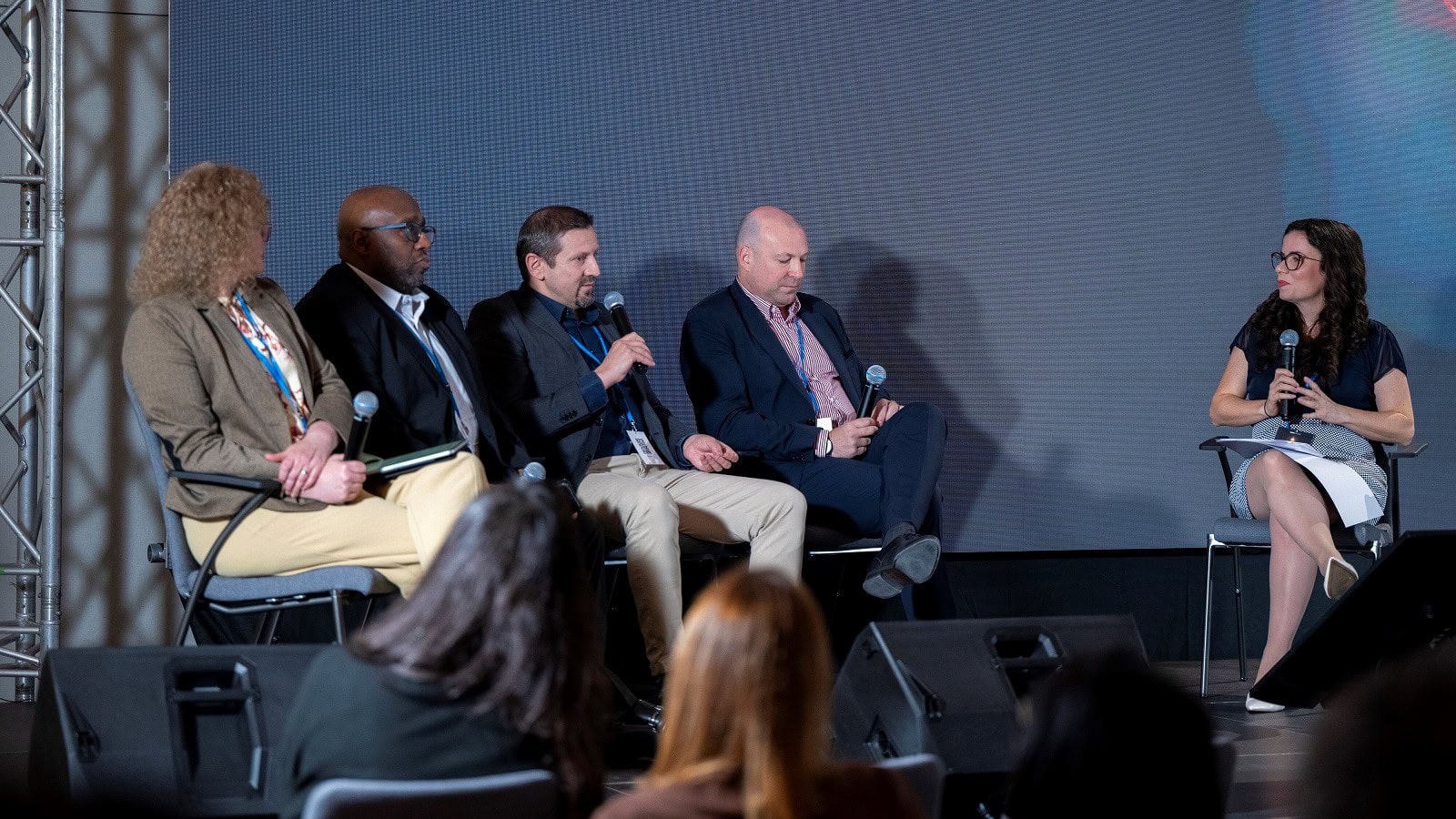 Vielfältiges Panel auf einer Konferenz, mit Diskussionsteilnehmern im Business-Format und einem Moderator, der Fragen stellt, vor einem großen digitalen Bildschirm, bei einer Medien- oder Bildungskonferenz.